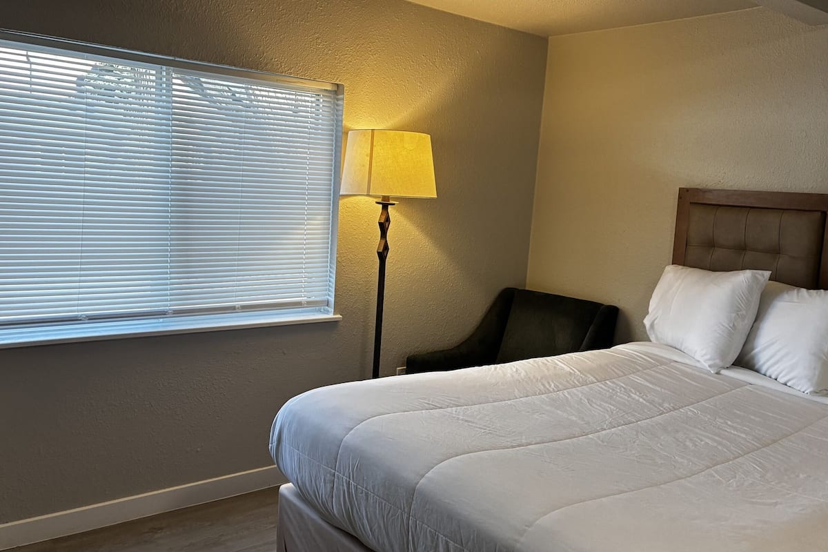 Una habitación de hotel con cama, sillón, lámpara y ventana con persianas.