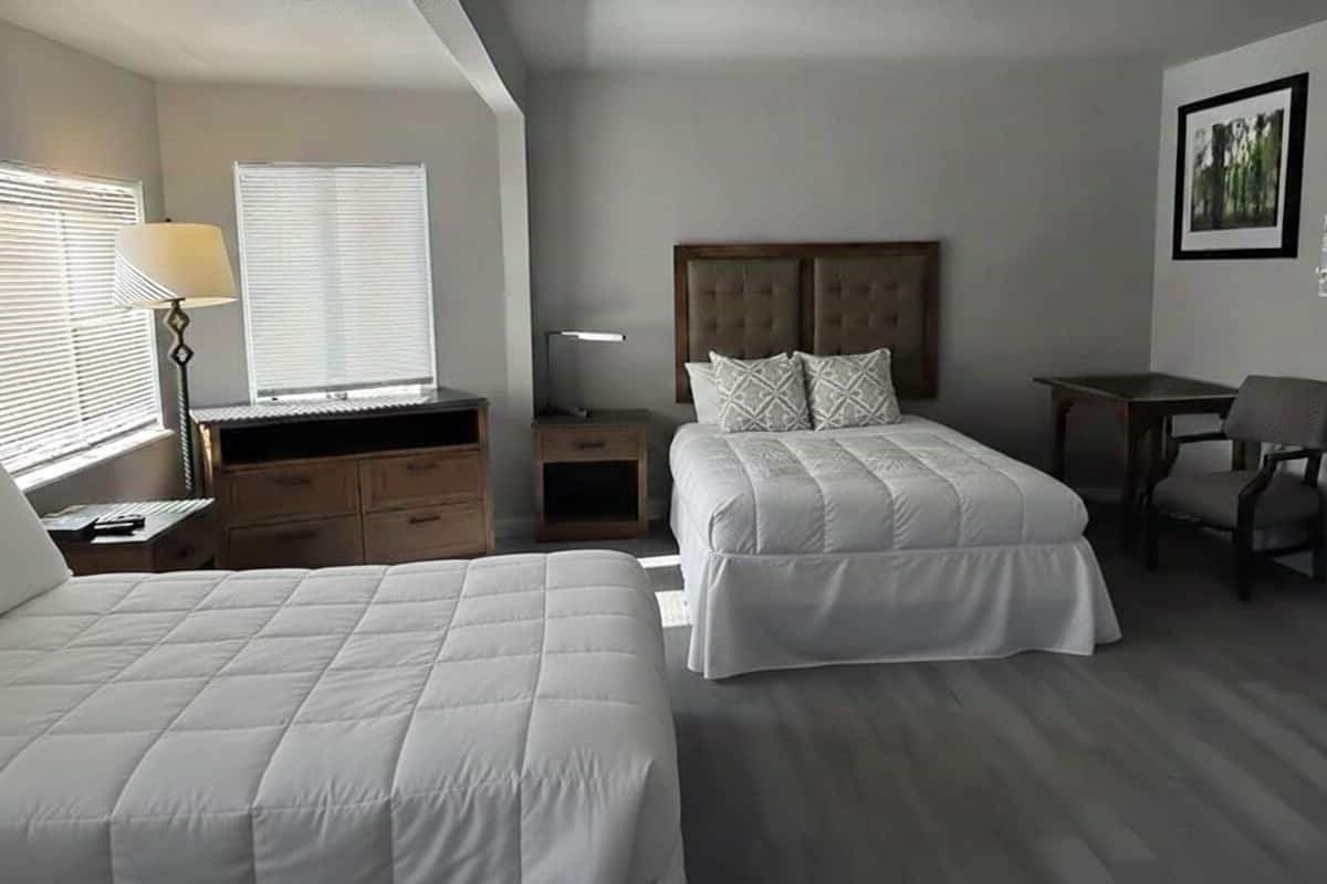 Quarto de hotel com duas camas, uma escrivaninha, uma cadeira, um abajur e um quadro na parede.