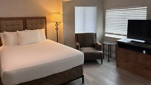 Habitación de hotel con una cama grande, televisión, una silla, lámpara y ventana con persianas.