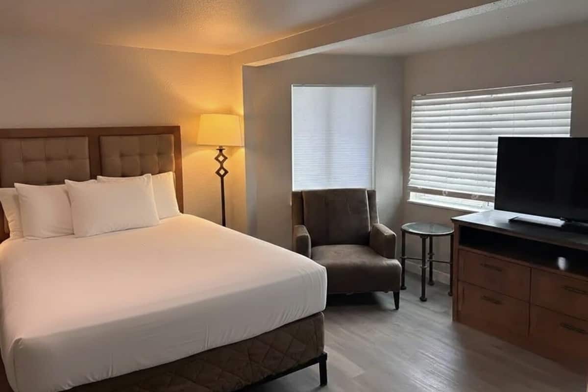 Habitación de hotel con una cama grande, televisión, una silla, lámpara y ventana con persianas.