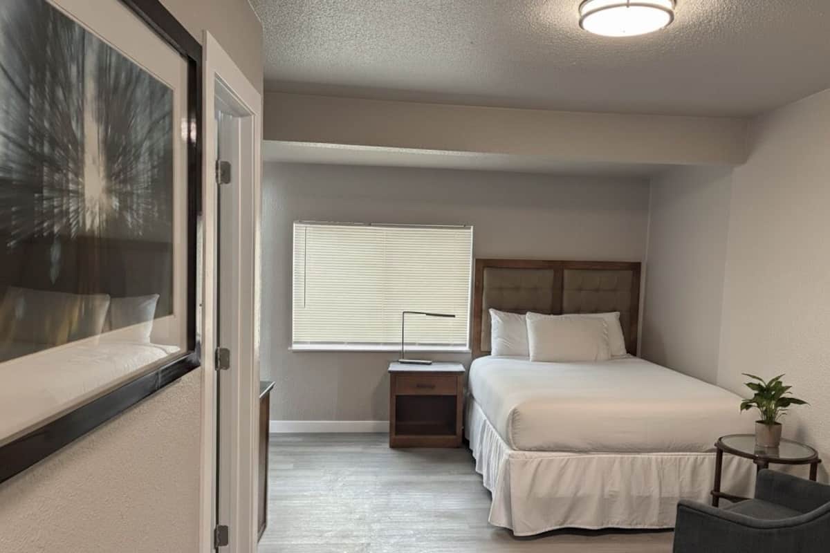 Quarto com cama, mesa de cabeceira, luminária, cadeira, janela e porta para outro cômodo.