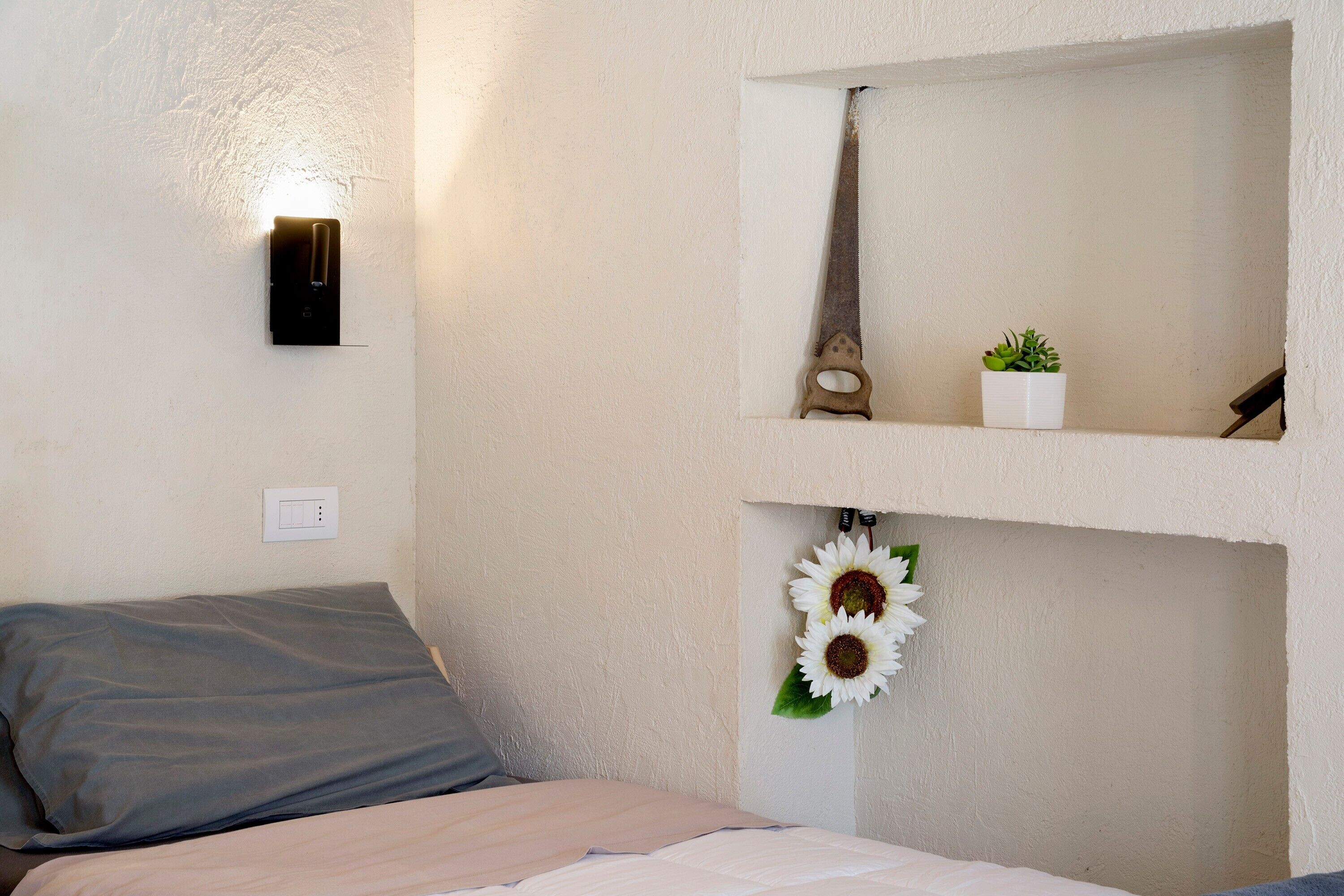 Une chambre à coucher comprenant un lit, une lampe fixée au mur, un miroir, une petite plante et un objet décoratif.