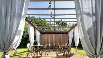 A covered outdoor seating area with tables and chairs, a wooden pergola, and a backdrop of trees.