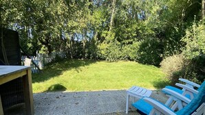 A patio area with a table and two blue chairs, surrounded by a grassy lawn and dense trees. - Nature Escape Home Near Veere (Gapinge)