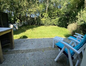 Eine Terrasse mit Tisch und zwei blauen Stühlen, umgeben von einer grünen Rasenfläche und dichter Baumgruppe.