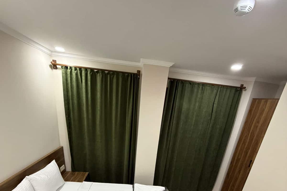 Una habitación con cama, mesita de noche, ventana con cortinas y una lámpara en el techo.
