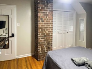 A bedroom with a brick accent wall, white closet doors, a bed with a grey blanket, and a flat-screen TV on a stand. - Staysobrio Luxury Stays (Gastonia)