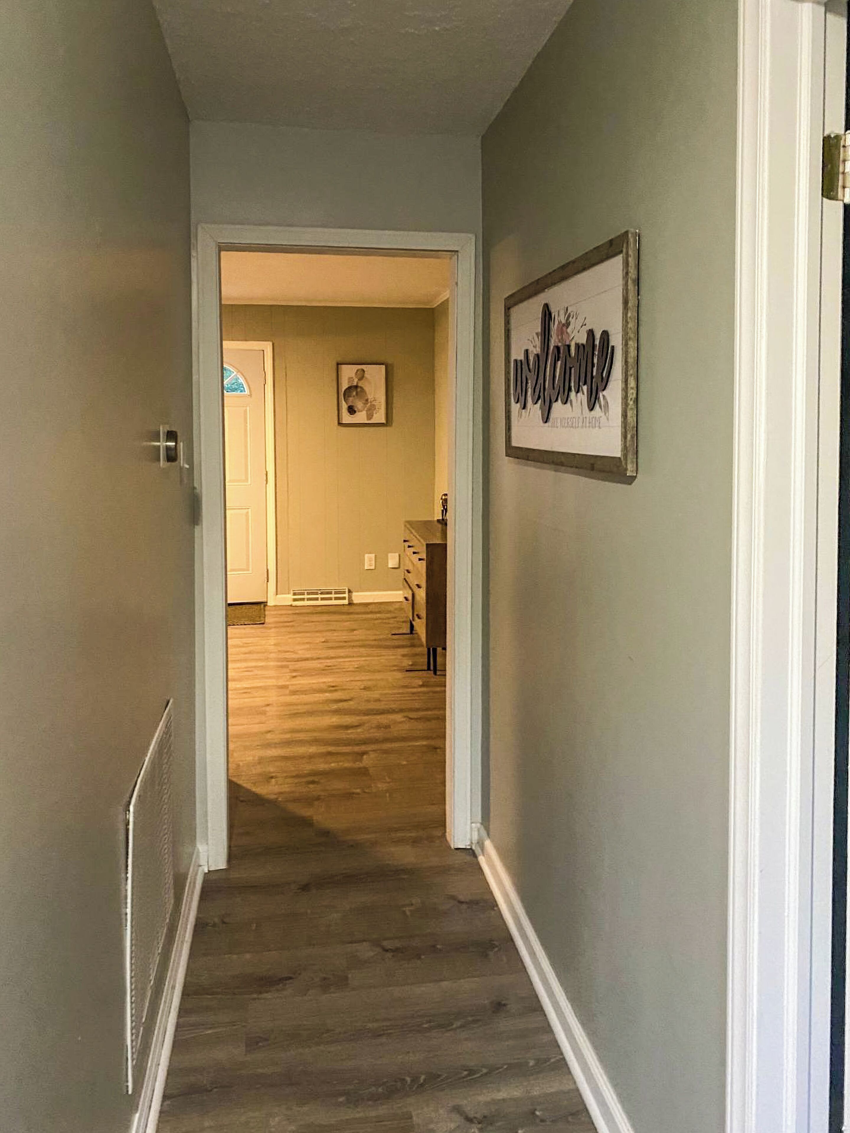 A hallway with a wooden floor leading to a room with a door and a framed welcome sign.