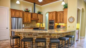 A kitchen with a central island, wooden cabinets, a stainless steel refrigerator, a built-in oven, and a stone countertop. - Highlife - Sleeps 23 - Frio River Access w/ Private Pool & Outdoor Kitchen (Concan)