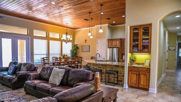 A spacious living room with a leather sofa, dining area, kitchen with wooden cabinets, and a high ceiling with exposed beams. - Highlife - Sleeps 23 - Frio River Access w/ Private Pool & Outdoor Kitchen (Concan)