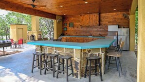 An outdoor kitchen with a stone island, bar stools, a grill, and a refrigerator. - Highlife - Sleeps 23 - Frio River Access w/ Private Pool & Outdoor Kitchen (Concan)