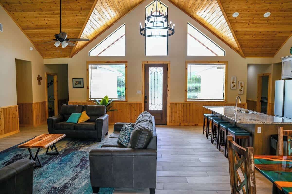 A spacious living area with a high ceiling, a large window, a sofa, a coffee table, and a kitchen area with a bar.