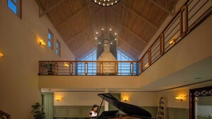 Un piano de cola en un amplio salón con piso a cuadros, vigas de madera y un balcón con muro de piedra.