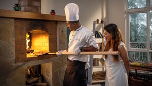 Un chef preparando comida en un horno de ladrillo, una estantería con libros y una ventana con vistas a árboles.