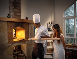 Un chef preparando comida en un horno de ladrillo, una estantería con libros y una ventana con vistas a árboles.