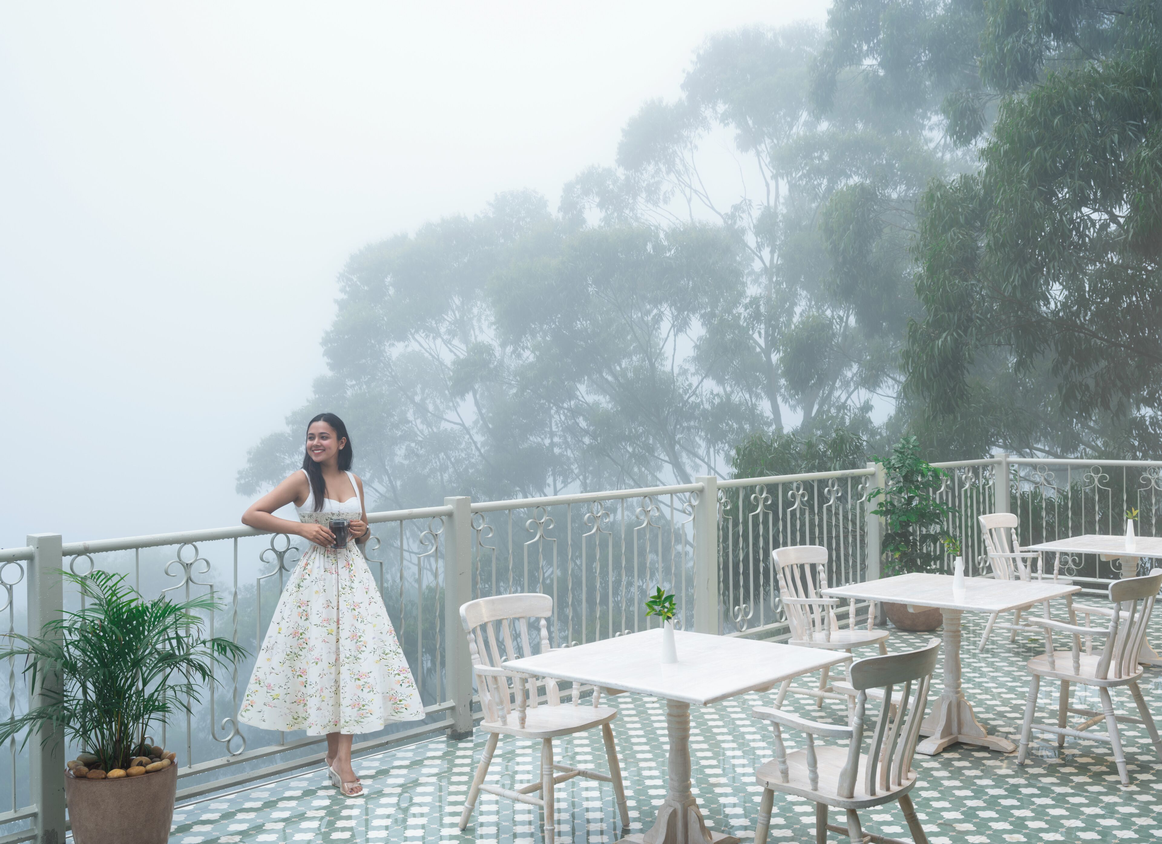 A woman standing on a balcony with a railing, overlooking a foggy landscape with trees.