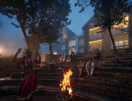 Una persona vestida con traje tradicional actuando cerca de un brasero, con un edificio moderno al fondo.