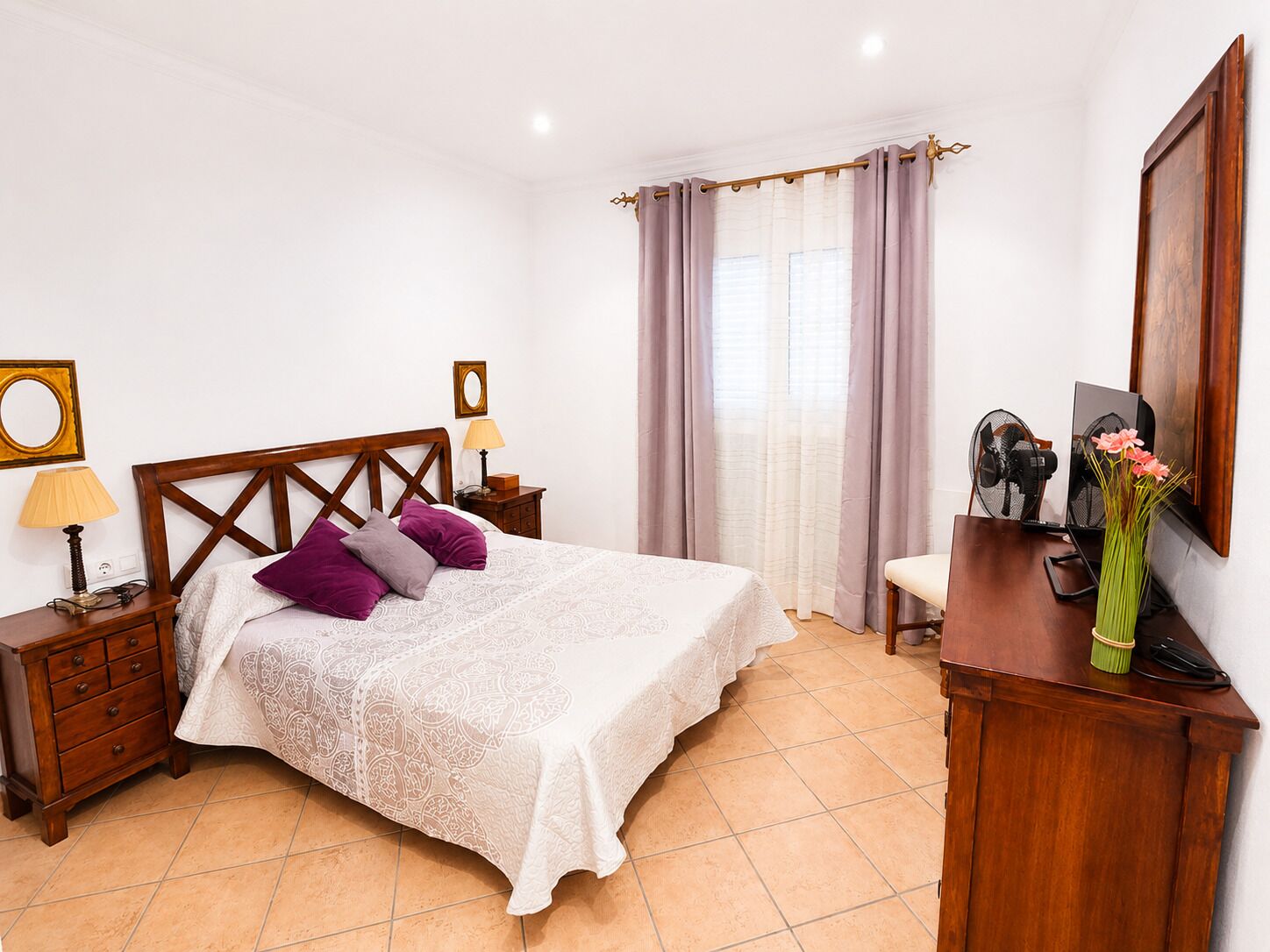 Ein Schlafzimmer mit einem hölzernen Bett, zwei Nachttischen, einem Schreibtisch mit Stuhl, einem Ventilator und einer Vase mit Blumen.