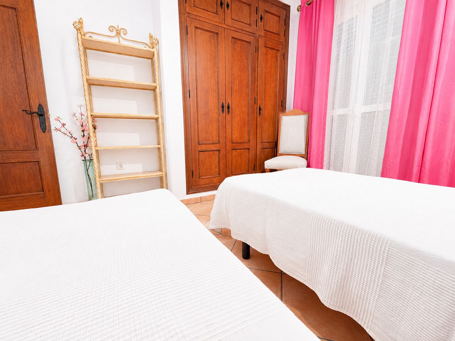 A bedroom with two beds, wooden wardrobe, and a window with pink curtains.