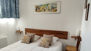 Une chambre à coucher comprenant un lit, des oreillers, une chaise, une table de chevet, une œuvre d’art accrochée au mur et une fenêtre avec des rideaux.