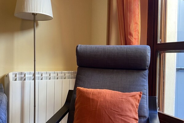 Una silla con cojín y almohada, un radiador, una lámpara y una ventana con cortinas.