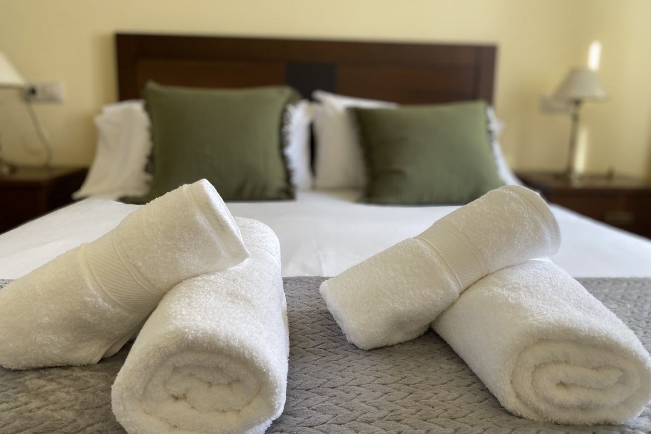A neatly made bed with white linens and rolled towels, a wooden headboard, and green pillows.