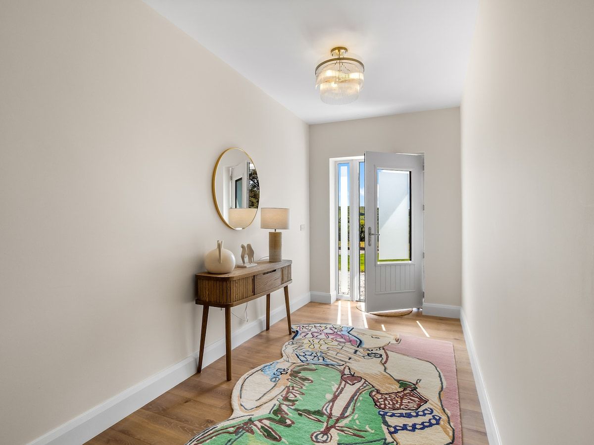 A hallway with a wooden floor, a round mirror, a small table with a lamp, and a colorful rug.