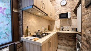 A compact kitchen with a microwave, stove, and washing machine, wooden cabinets, and a brick wall backsplash.