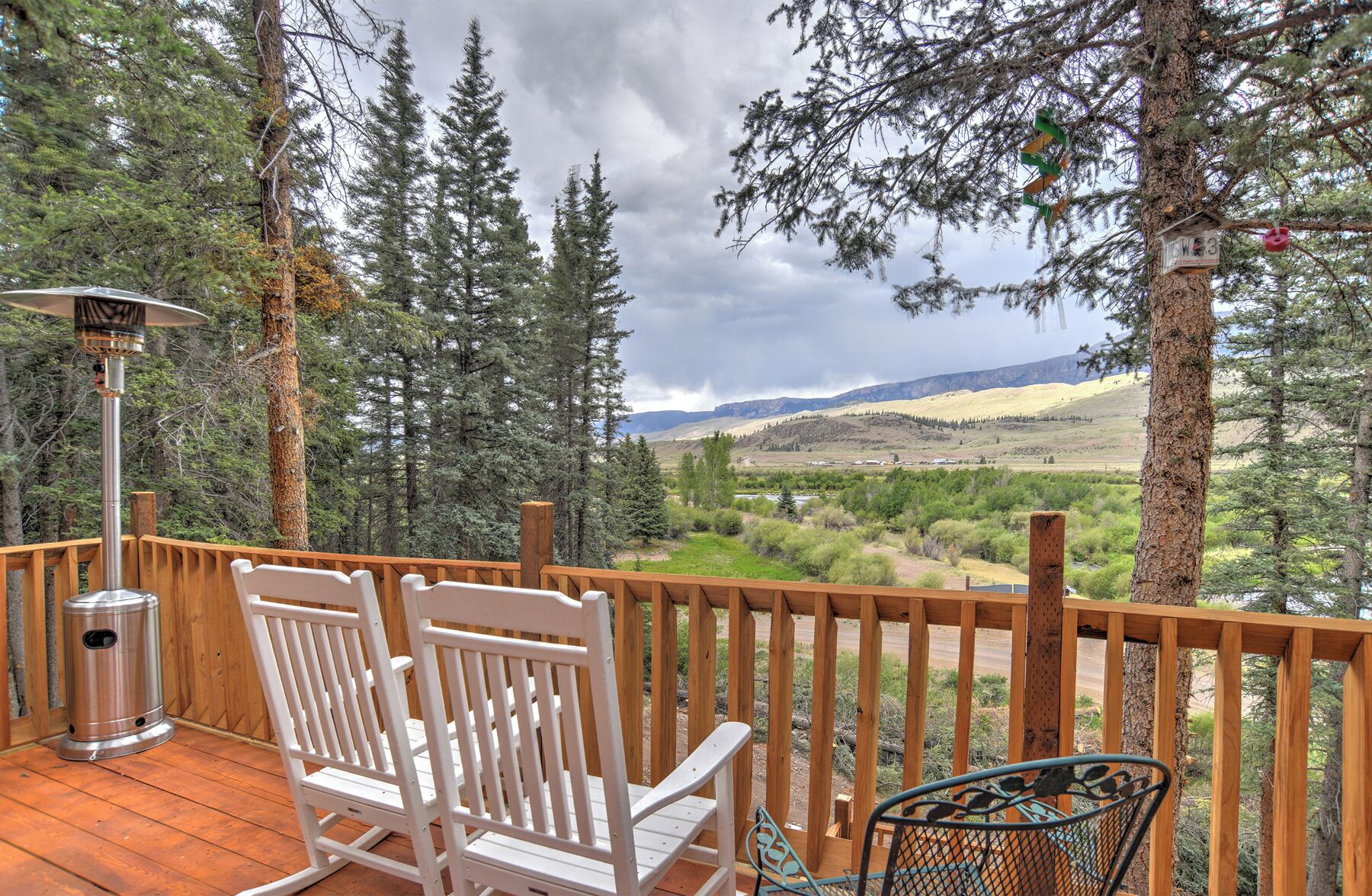 Une terrasse en bois avec des chaises blanches et une chaise en métal, donnant sur un paysage montagneux parsemé d’arbres.