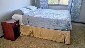 Un dormitorio con cama, mesita de noche, ventana con cortinas y piso alfombrado.