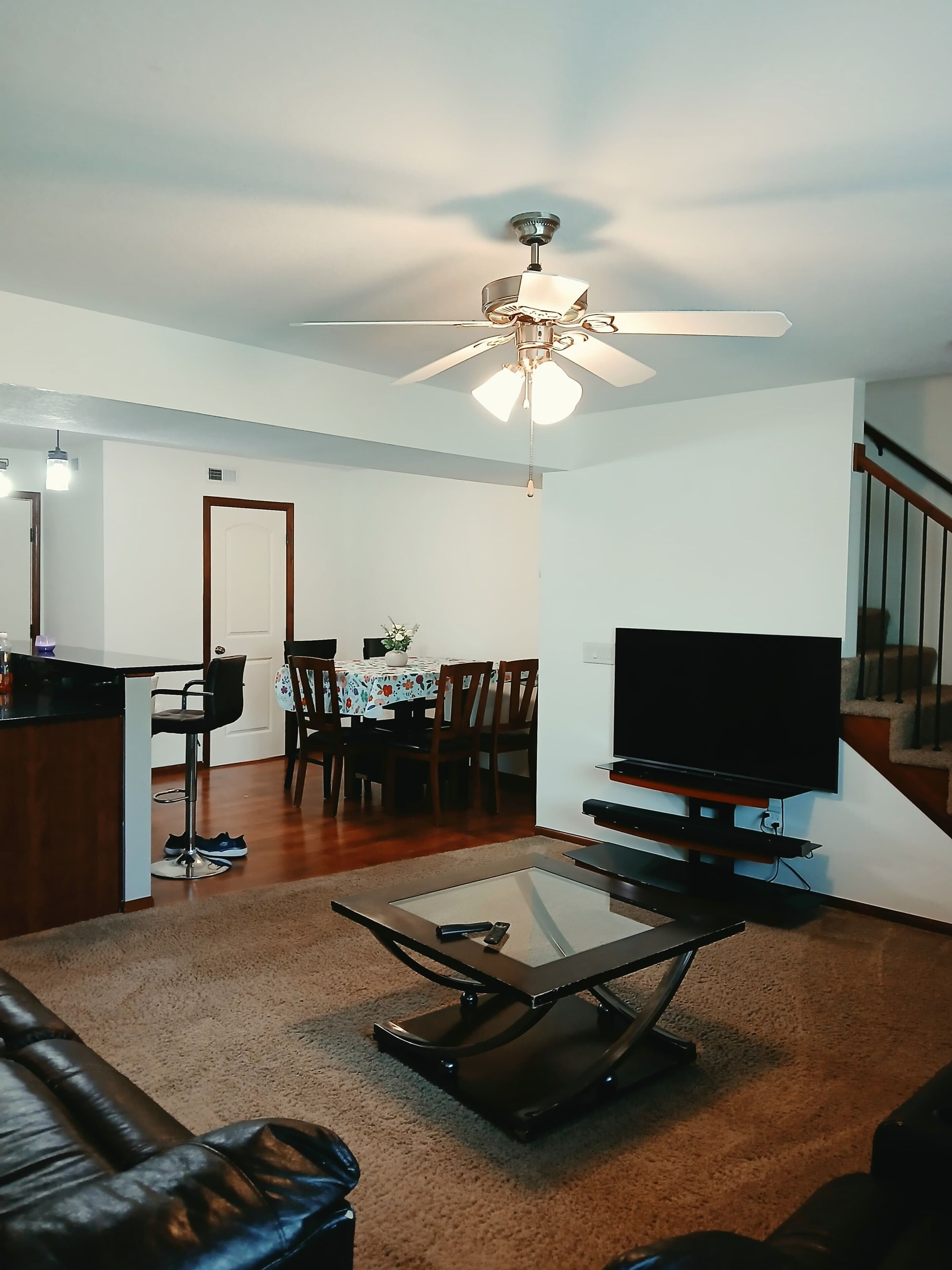 A living room with a ceiling fan, a television, a glass coffee table, a leather sofa, and a dining area with a table and chairs.
