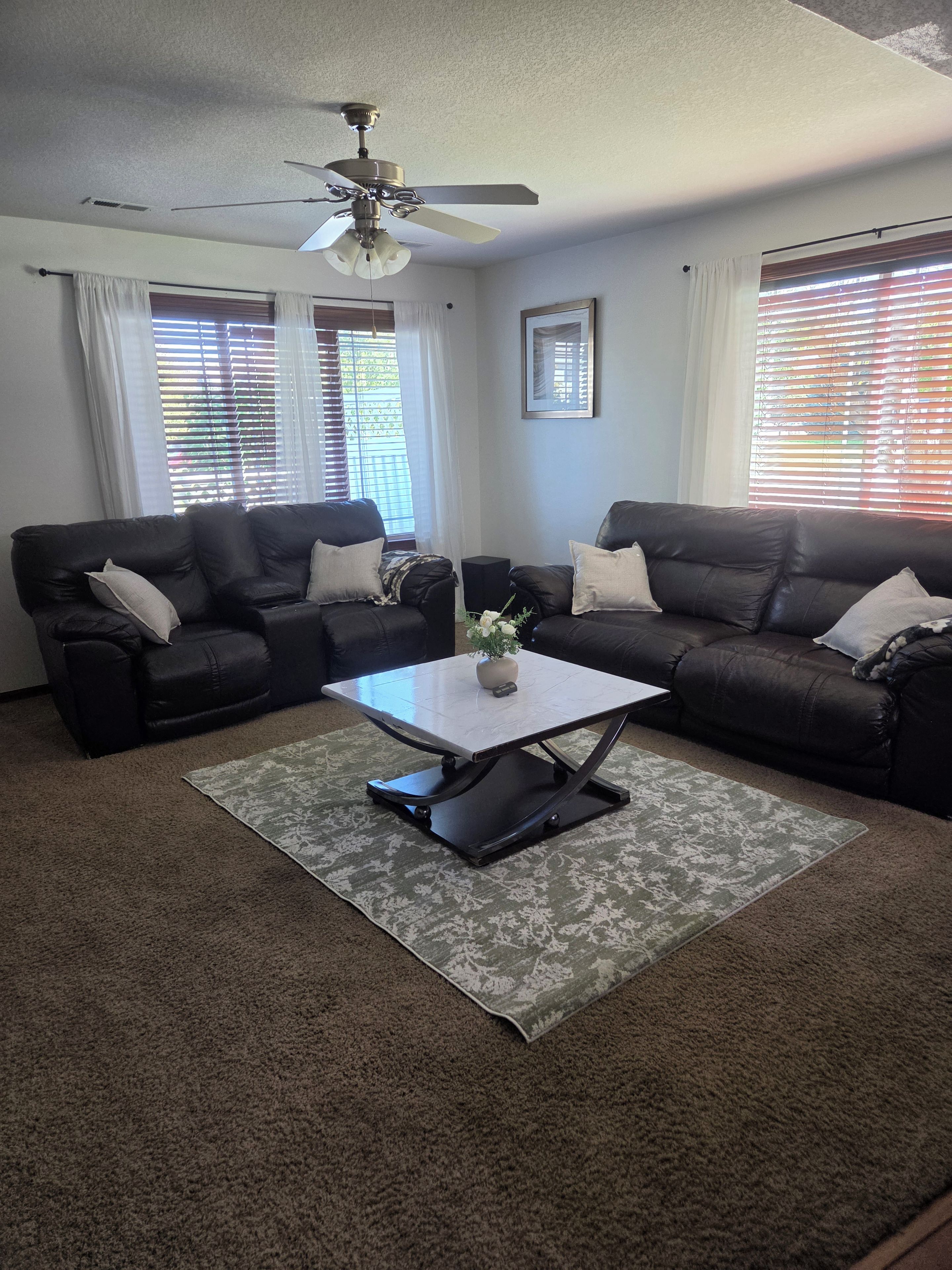 A living room with two sofas, a coffee table, a ceiling fan, and a window with blinds.