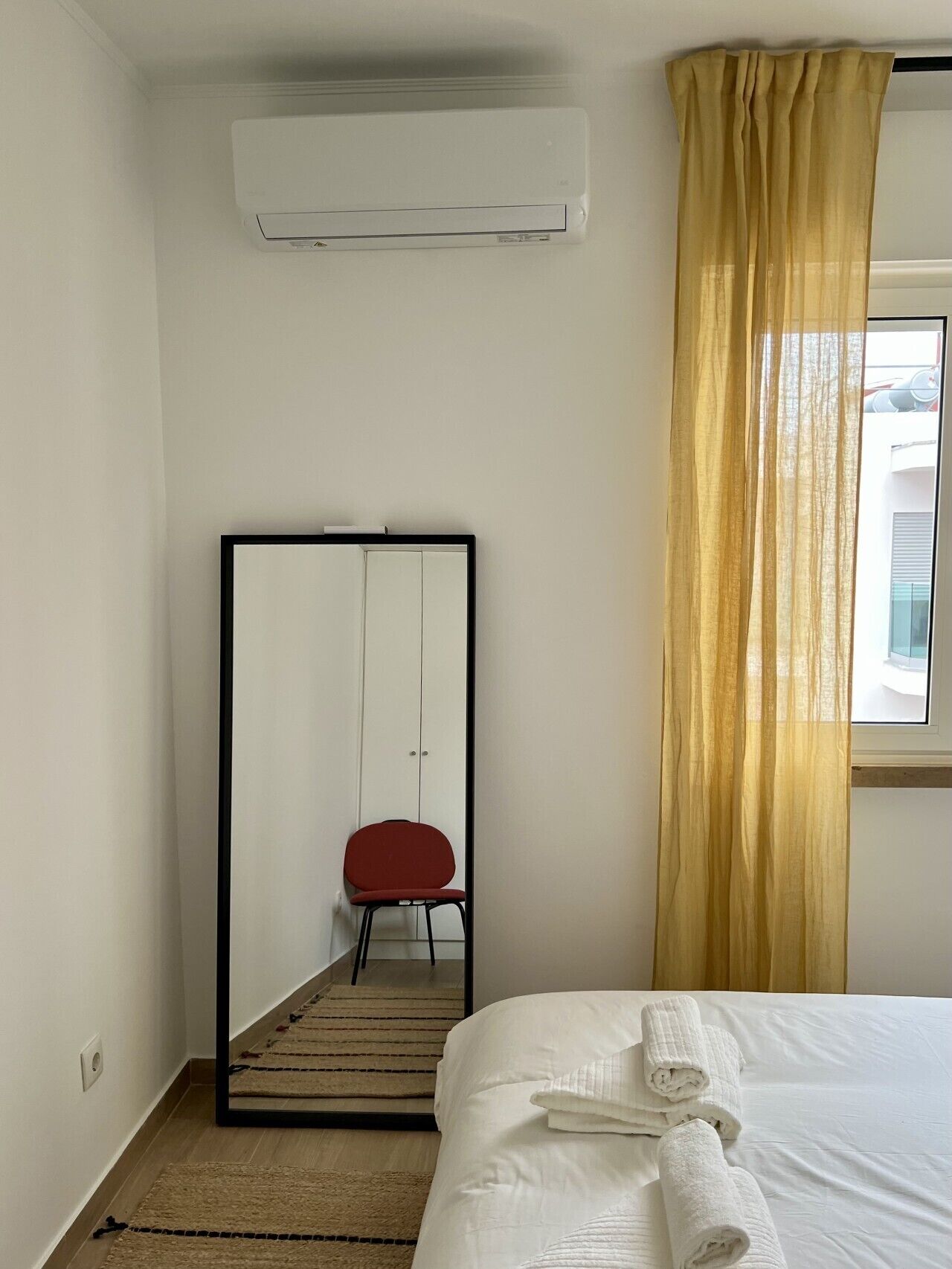Hotellihuone, jossa on sänky, punainen tuoli, liukuovi, ilmastointilaite ja ikkuna, jossa on verhot.