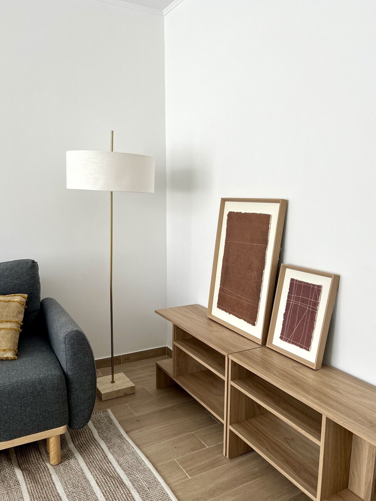 Un salon moderne avec un canapé gris, une lampe sur pied et deux œuvres d’art encadrées posées sur une console en bois.