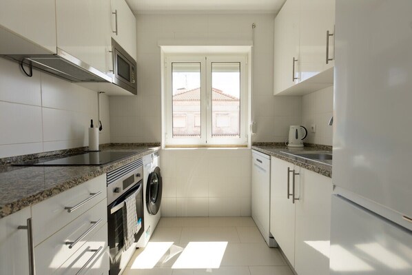 A modern kitchen with white cabinets, a built-in microwave, a washing machine, and a window providing natural light. - Modern Haven in Amadora (Amadora)