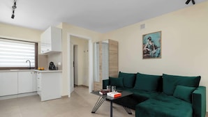 Ein modernes Wohnzimmer mit einem grünen Sofa, einem kleinen Couchtisch und einer Küchenzeile mit weißen Schränken und einem Fenster mit Jalousien.
