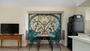 Ein Essbereich mit einem Glastisch, stuhlfarbenen Sesseln, einem Fernseher auf einem Holztisch und einem Kühlschrank.