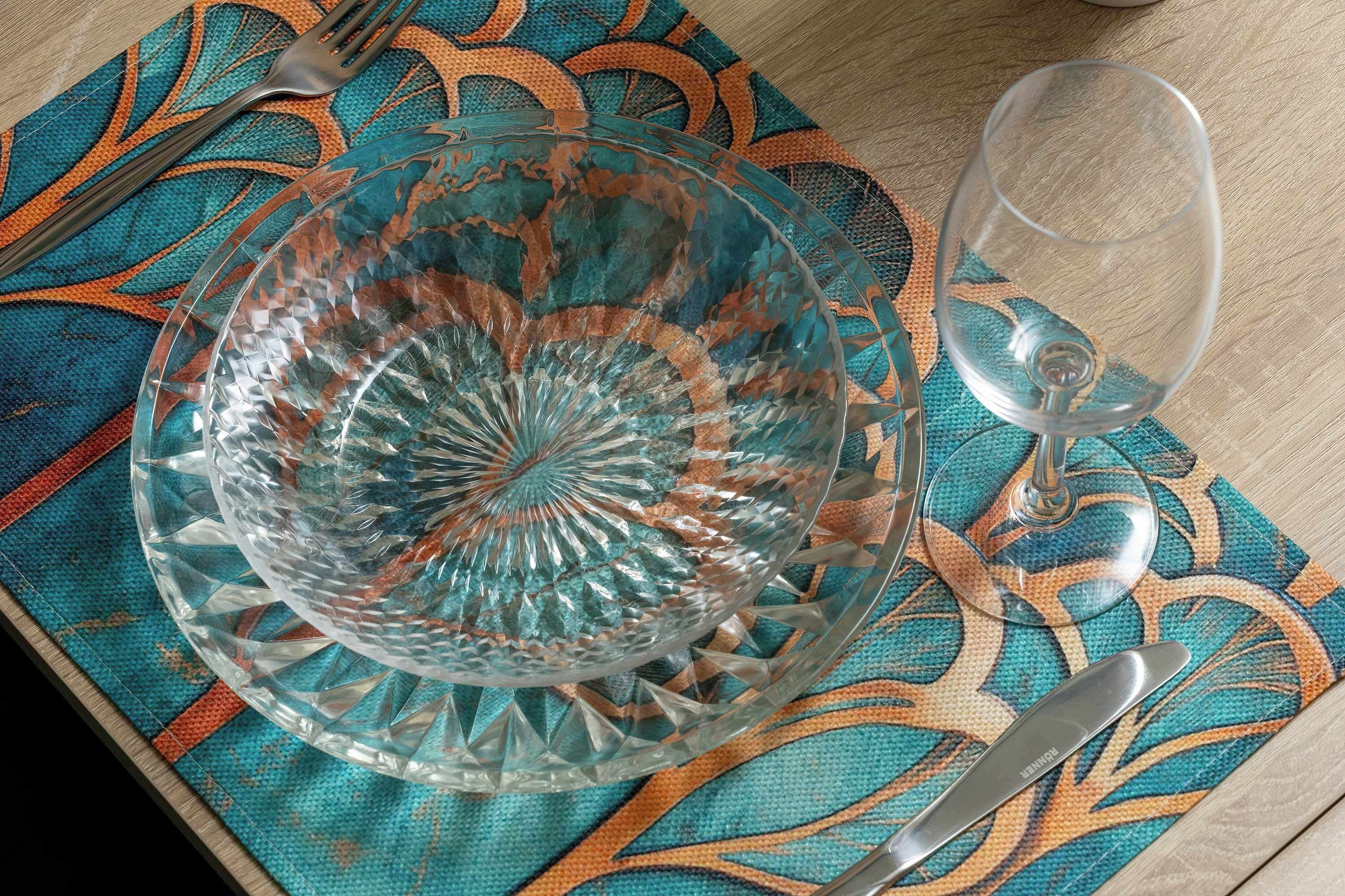 Une table dressée avec une assiette en verre transparent, un verre à vin et des couverts posés sur une nappe à motifs.