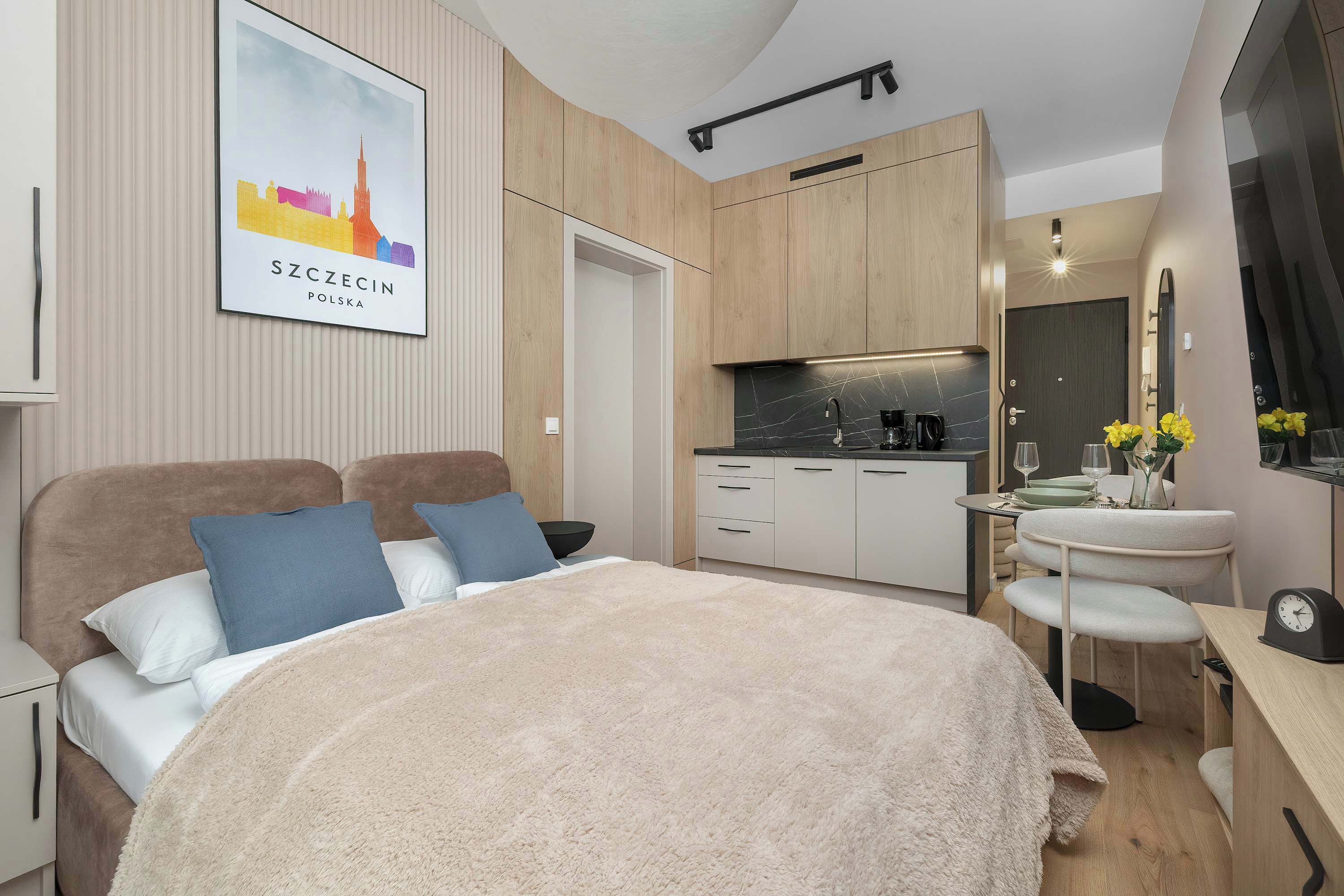 A modern hotel room with a bed, a small kitchenette, a bedside table, and a mirror.
