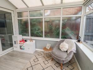 Ein Wintergarten mit großem Fenster, ein weißes Küchenschränkchen mit Geschirr, ein grauer Sessel und ein kleiner Tisch mit Bilderrahmen.