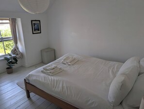 A bedroom with a bed, a window with curtains, a framed picture on the wall, and a potted plant. - Fairsands (Redruth)