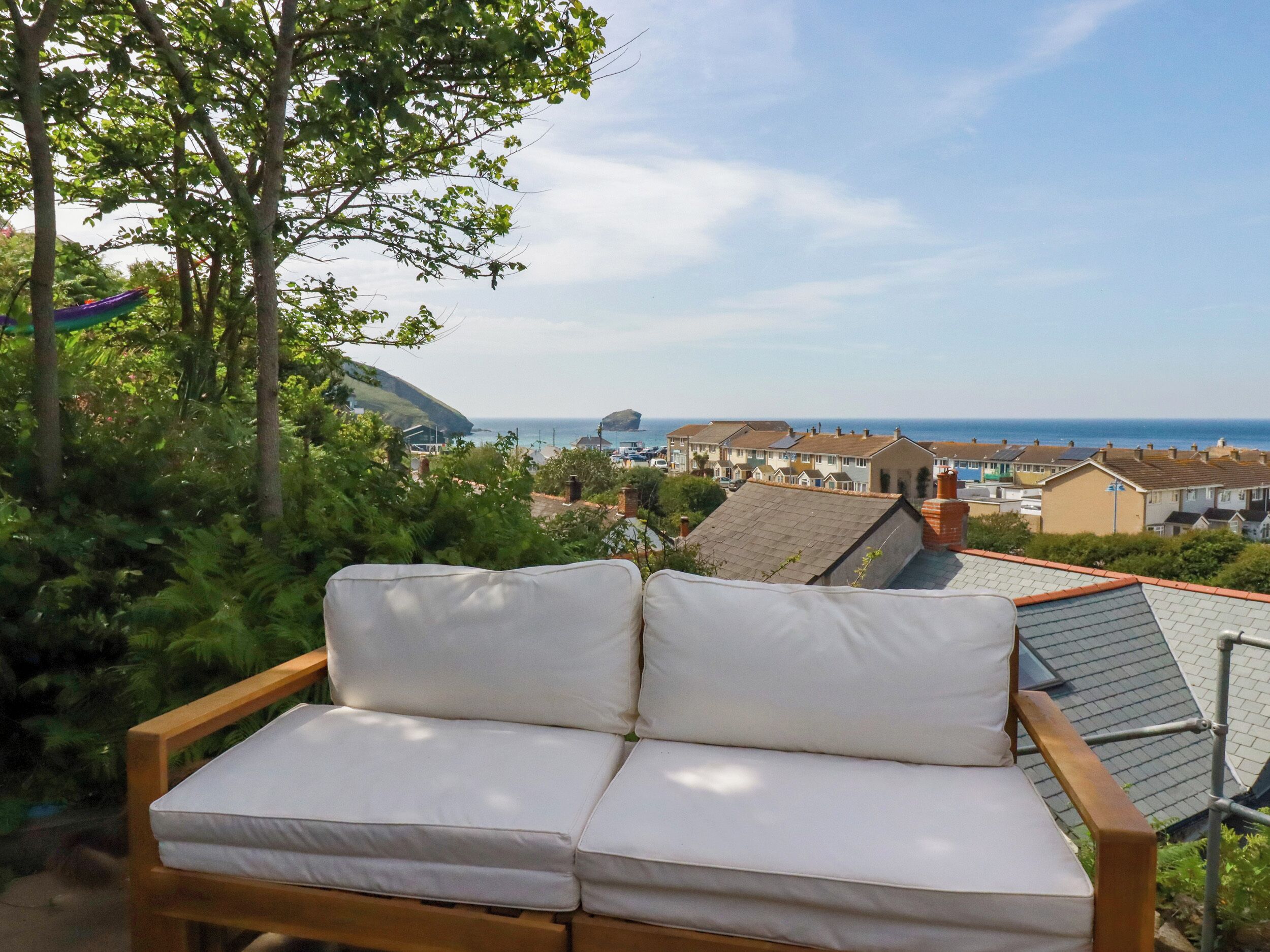 A wooden bench with white cushions, overlooking a coastal town with houses and a view of the sea.