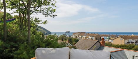 A wooden bench with white cushions, overlooking a coastal town with houses and a view of the sea.