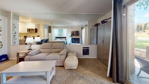 Ein modernes Wohnzimmer mit einer Couch, einem Hocker und einem Esstisch. Im Hintergrund ist eine Küche mit einer Mikrowelle und Schränken zu sehen.