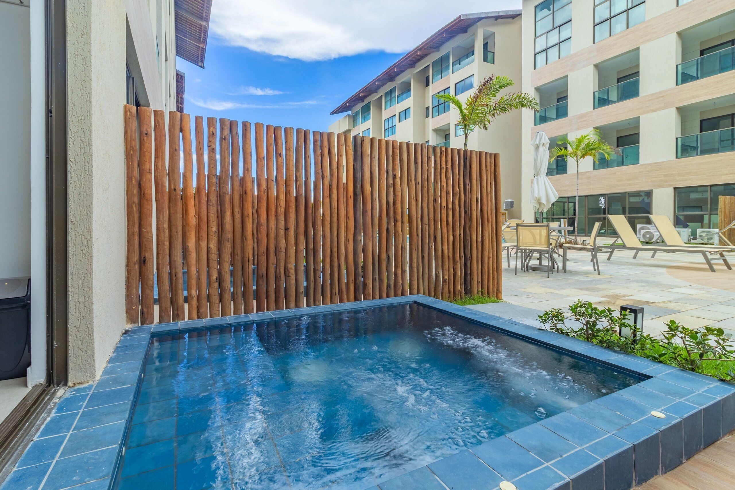 Piscina com cerca de madeira, área de terraço com cadeiras, e um edifício com vários andares e varandas.