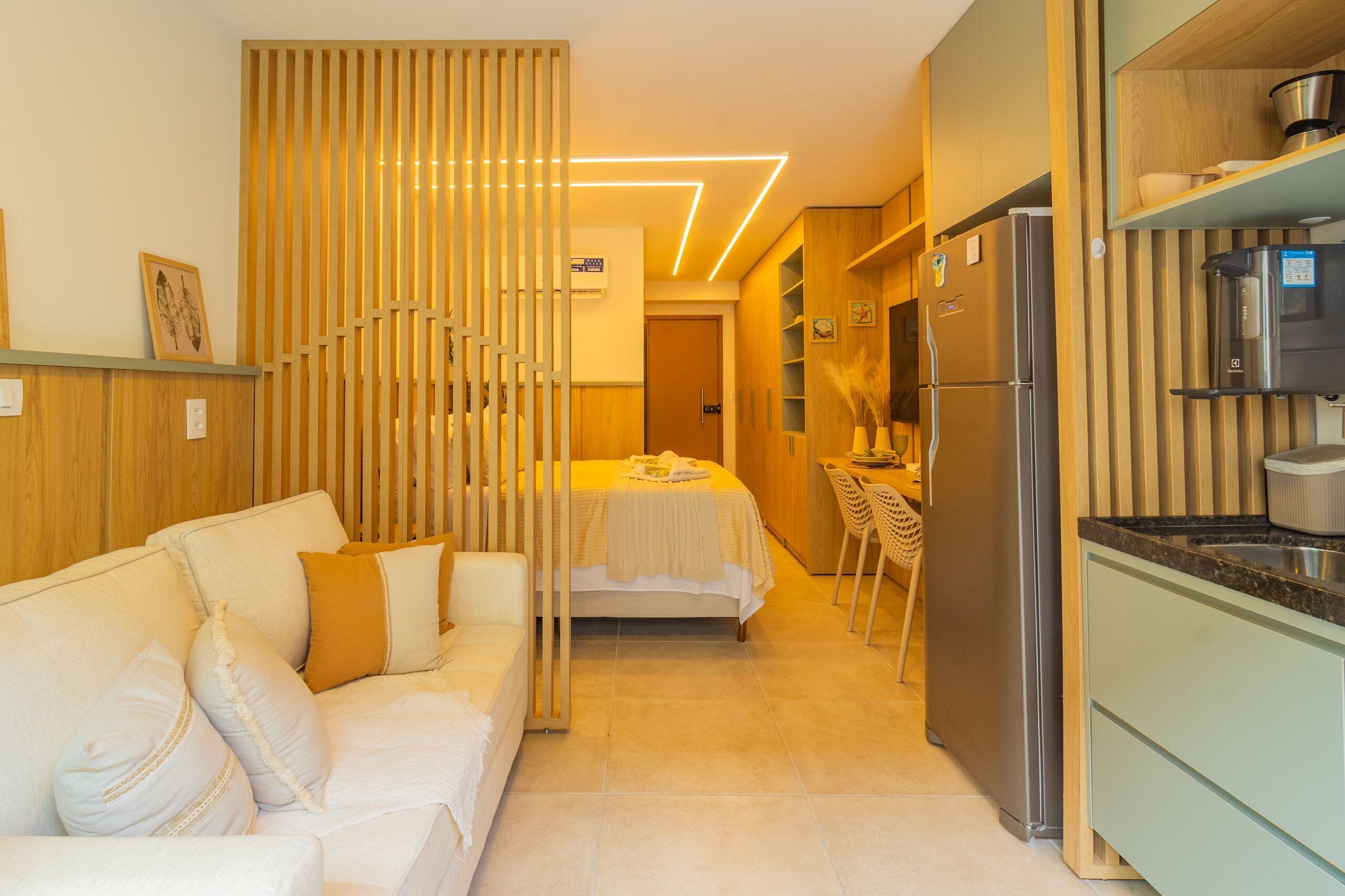 A modern kitchen with a refrigerator, a dining area with a table and chairs, and a living area with a sofa and cushions.