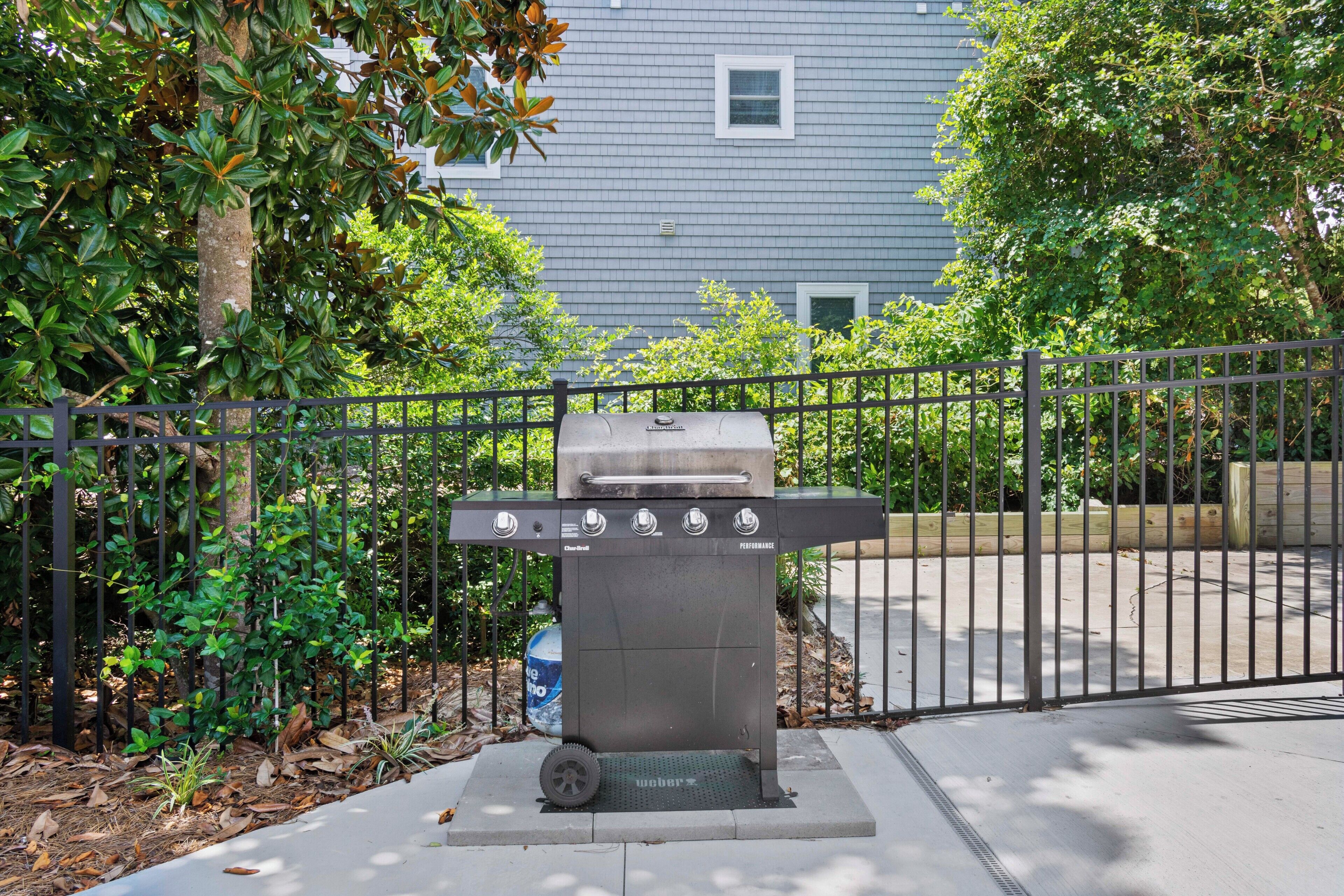 A Weber gas grill with four burners, a side shelf, and a footrest, set against a fence with a building in the background.