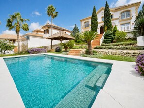 A swimming pool with a clear blue water, surrounded by a well-maintained garden with various plants and a large house with a tiled roof in the background. - Frontline Golf Villa in the stunning La Cala Golf. Perfect for families and golfers! (Mijas)