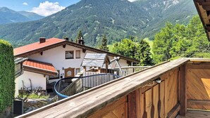 Ein hölzerner Balkon mit Blick auf eine gebirgige Landschaft, mit einem traditionellen Gebäude mit rotem Dach und einer Holzdielen-Terrasse.