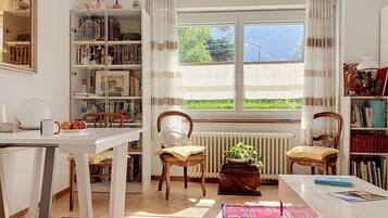 Une pièce avec une grande fenêtre, une bibliothèque, un radiateur et une table avec des chaises.